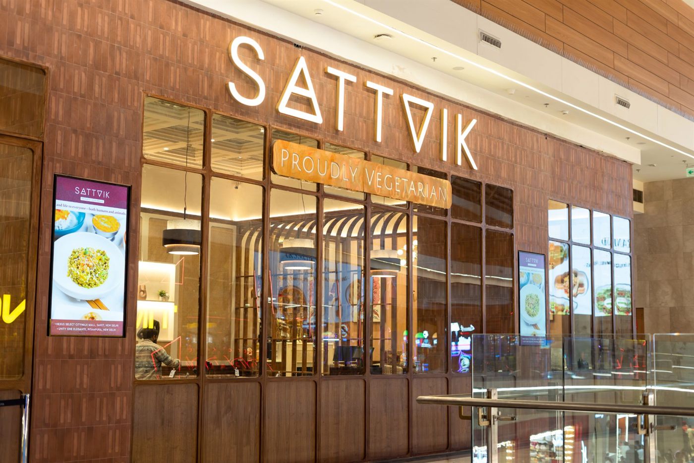 The Sattvik storefront in the mall, with its lit sign, the Proudly Vegetarian panel and a glass front looking into the dining room